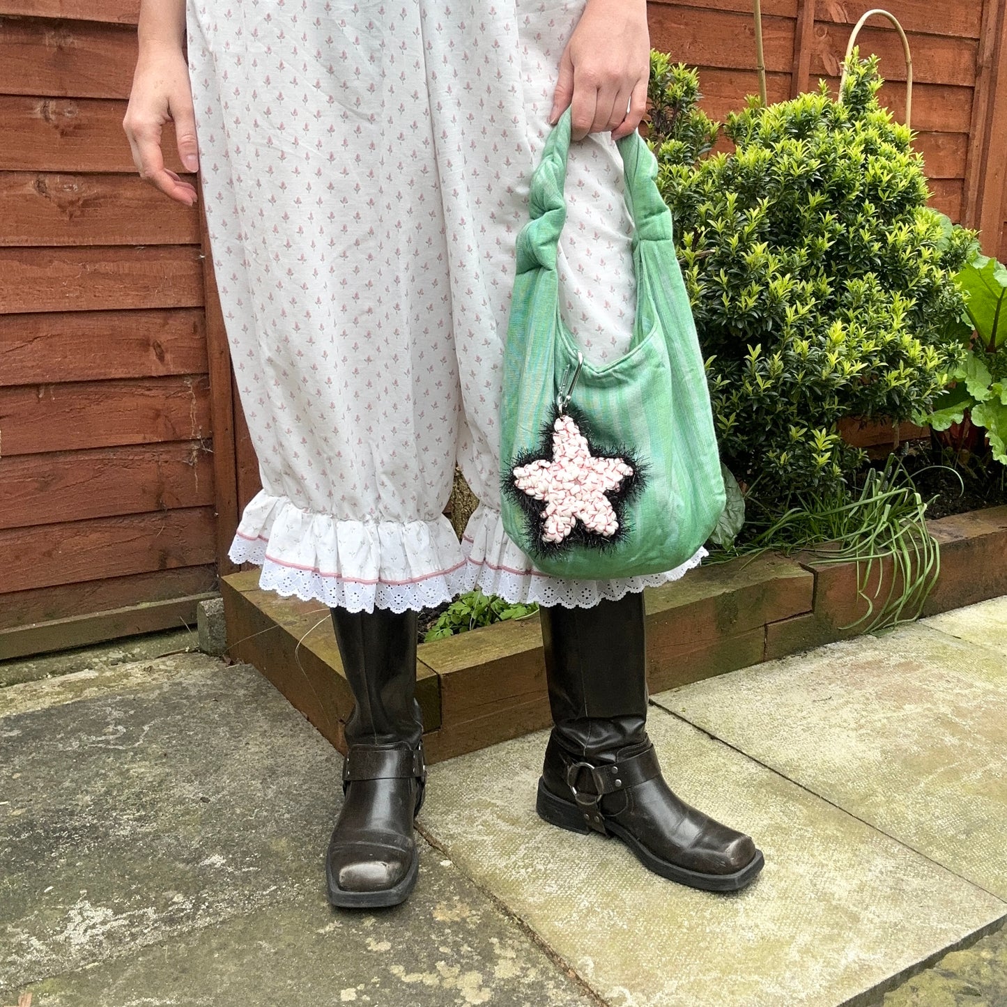 Model holding a green handbag with a Sloe Mosey star bag charm, wearing a white dress and black boots, standing outdoors.